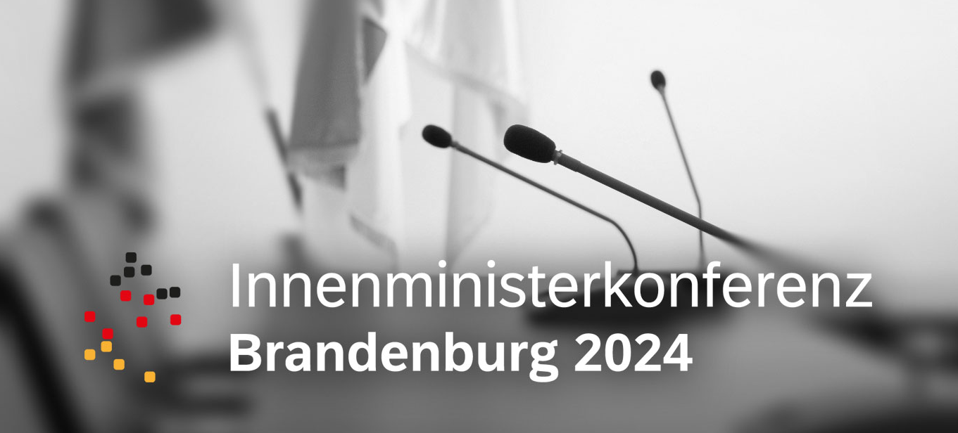 Foto von einem Beratungstsich mit Tischmikrofonen und dem Logo der IMK Brandenburg 2024 im Vordergrund